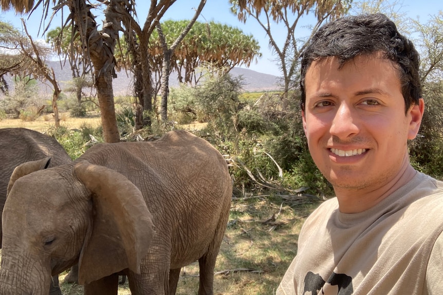 A man in a tan t-shirt takes a selfie in front of two elephants under some trees of an African plain with mountains in the back