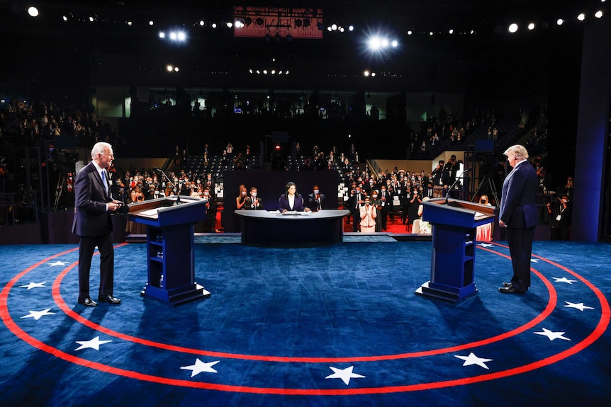 Joe Biden and Donald Trump stand far apart on a blue stage in front of a masked audience.