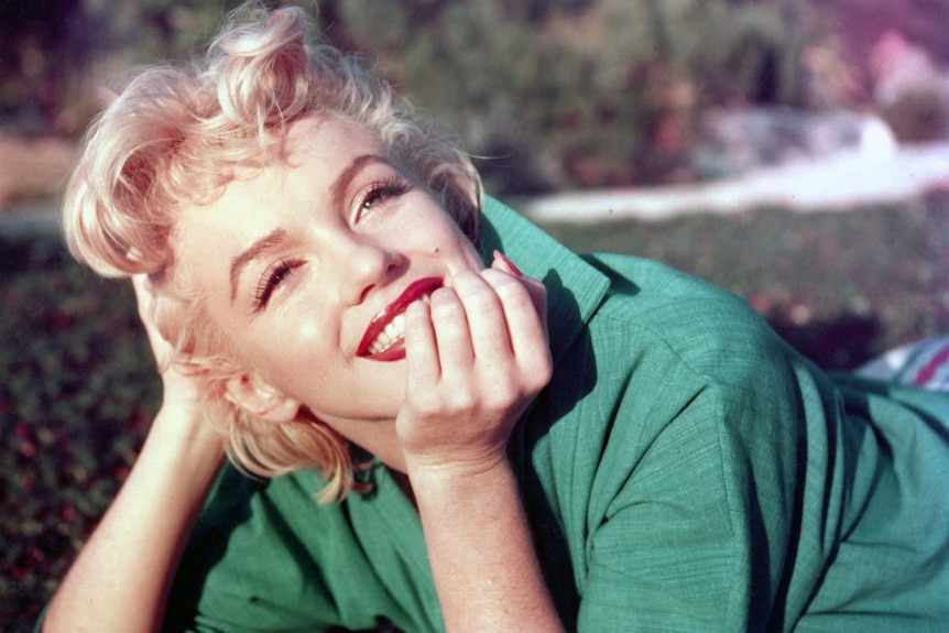 Marilyn Monroe lays on her stomach on a lawn, smiling into the distance as she rests her chin on her right hand