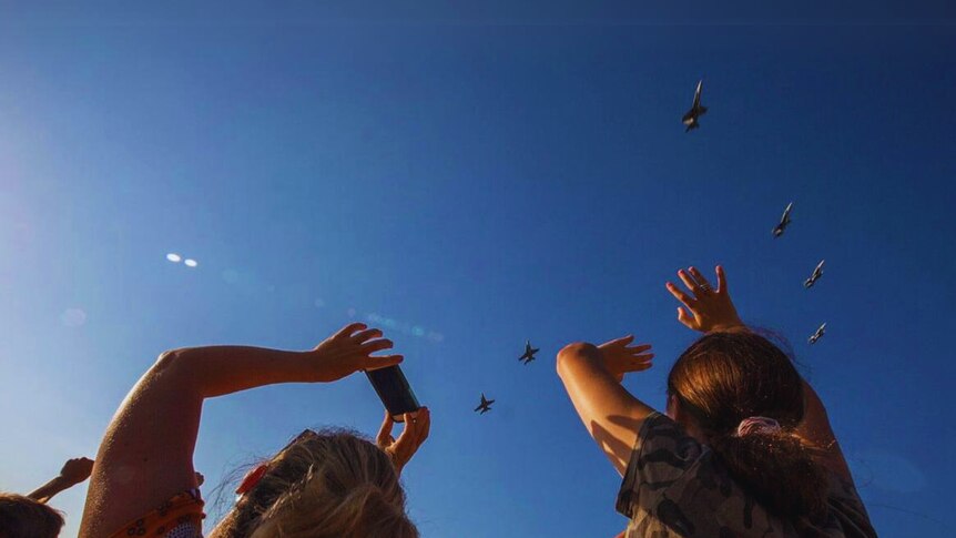 Several fighter jets in the sky seen from below and behind a couple of people looking up.