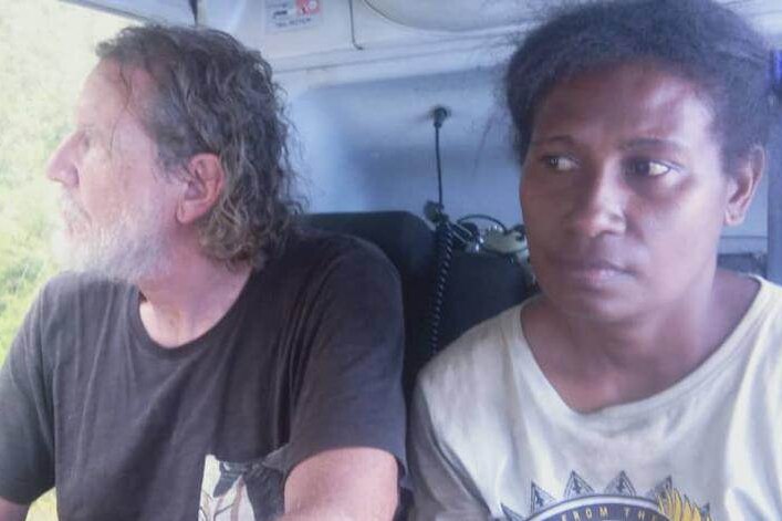 A man and a woman pictured inside a helicopter, both looking out the door at the green forest below