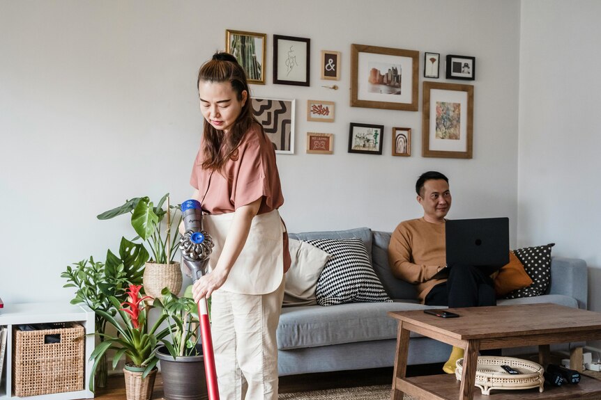 A woman vacuuming the floors while a man sits on a couch and uses a laptop. The living room has plants and prints on the wall. 