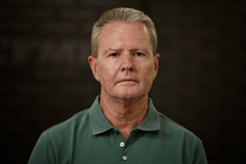 A man standing inside a room in front of a black wall, looking serious.