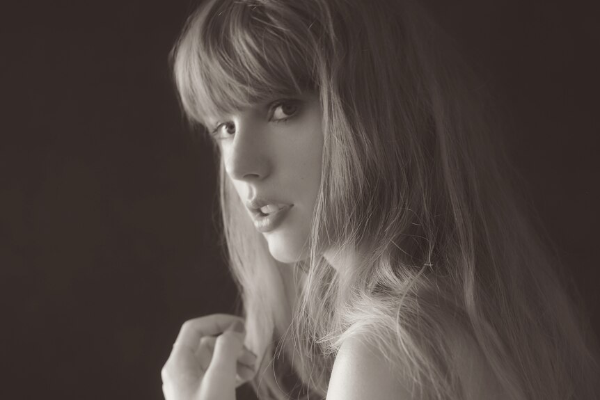 Taylor Swift in a black and white image, not smiling, looking over her shoulder, long blonde hair