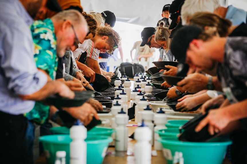 Group of people in colourful t-shirts lined up at tables panning for gold in big plates