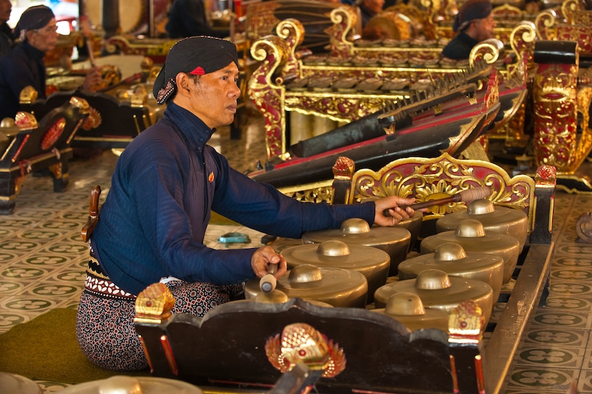 A close up of a Javanese bonang player sitting on the ground, surrounded by other gamelan players. 