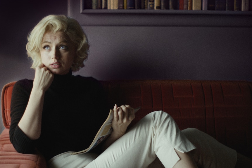 White woman with curly blonde hair in black turtle-neck and cream pants holds book while sitting on red couch.