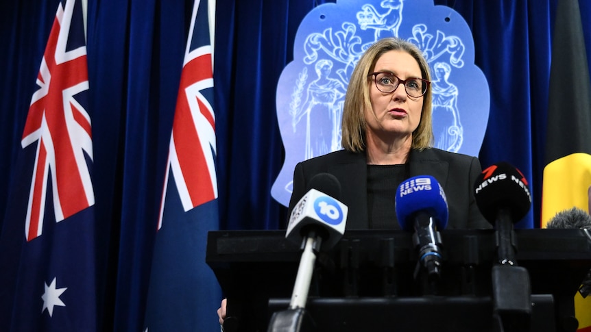 Jacinta Allan stands at a lectern with microphones in front of her. Behind her is the Australian flag