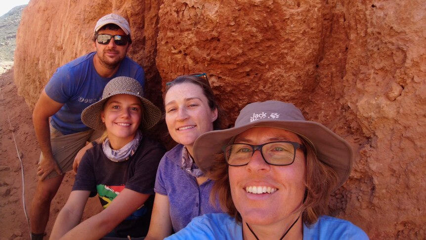 Four people smile in front of a termite mound.