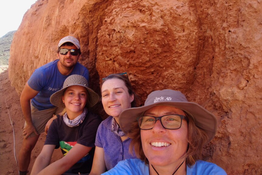 Four people smile in front of a termite mound.