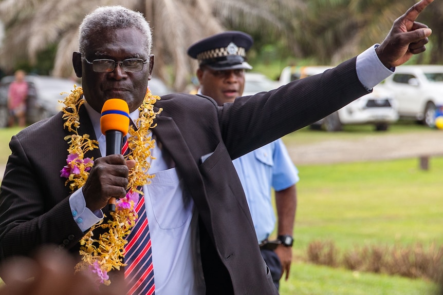 A man wearing a suit and a flower garland around his neck holds a microphone and points upwards.