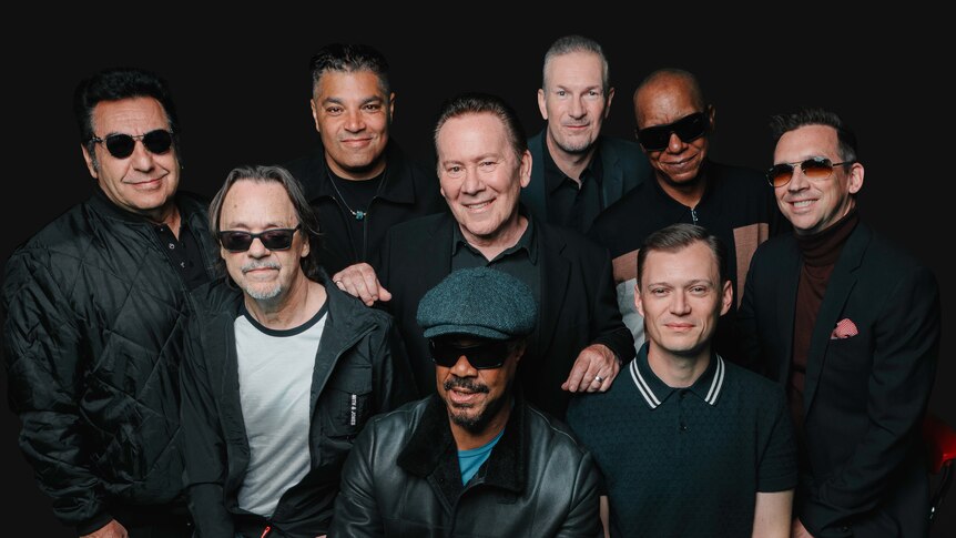Black background with nine men from waist up huddled together smiling at camera. 