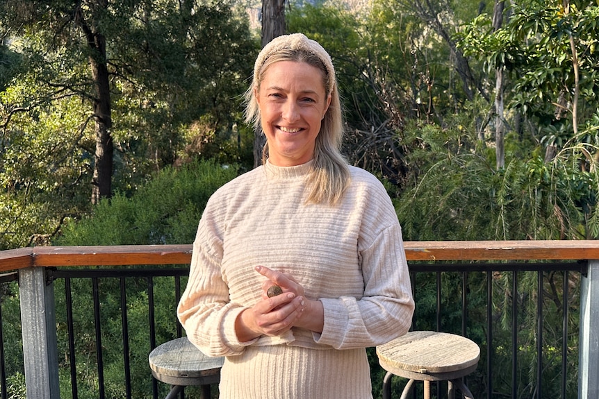 A woman, Lisa Whyte, standing on her balcony with trees, a fence and barstools in the background