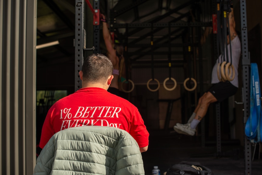 A young man sits on a chair waching athletes do pull ups and other exercises.