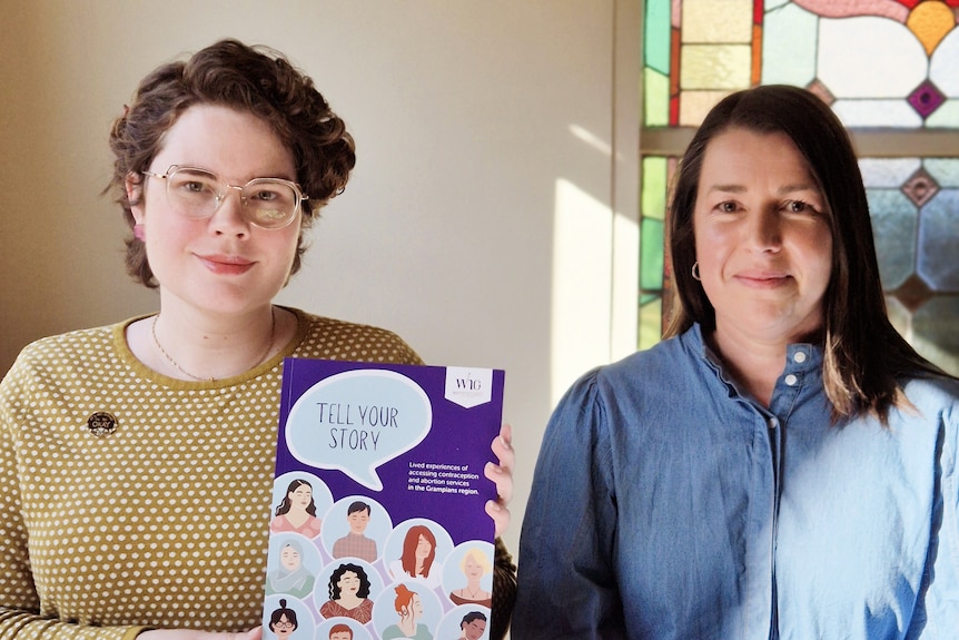 Two women at a table  holding up a purple document titled Tell Your Story, which features illustrations of women on the front.