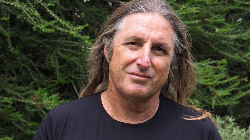 A man with long hair standing in front of green foilage smiling