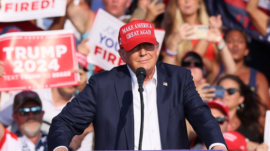 Donald Trump smiles while standing at a microphone with a crowd behind him.