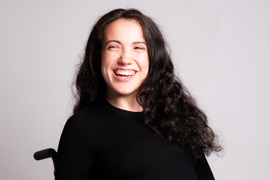 Hannah Diviney with long brown hair and a bright smile sitting in a wheelchair wearing all-black clothing