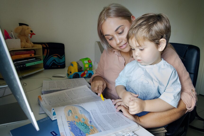 A woman sitting at a desk and looking at a textbook, with a small boy on her lap.