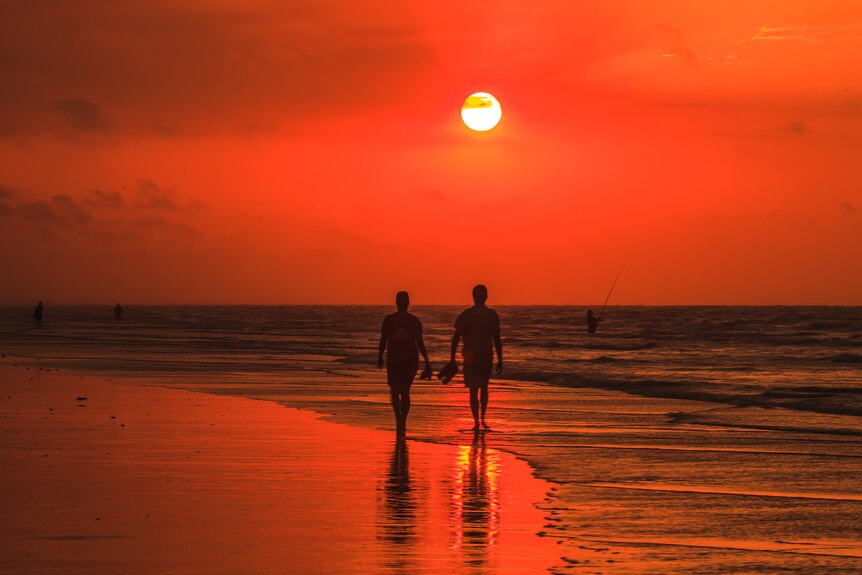 A silhouette of a couple walking on a beach, everything is red at sunset, people fishing in the background