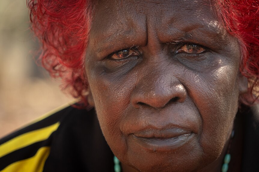 A middle-aged Indigenous woman with dyed red hair looks at the camera with a neutral expression.