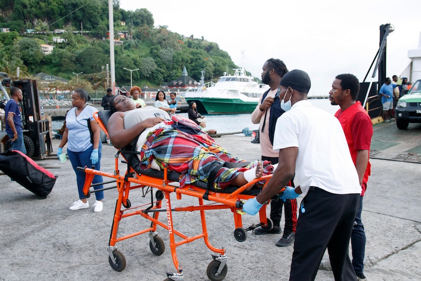 A woman lies on an orange stretcher, as a man in a white shirt pushes it across concrete.