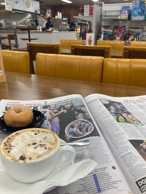 A donut is seen on a plate with a coffee near it, on top of a newspaper open to the sports section at a cafe table.