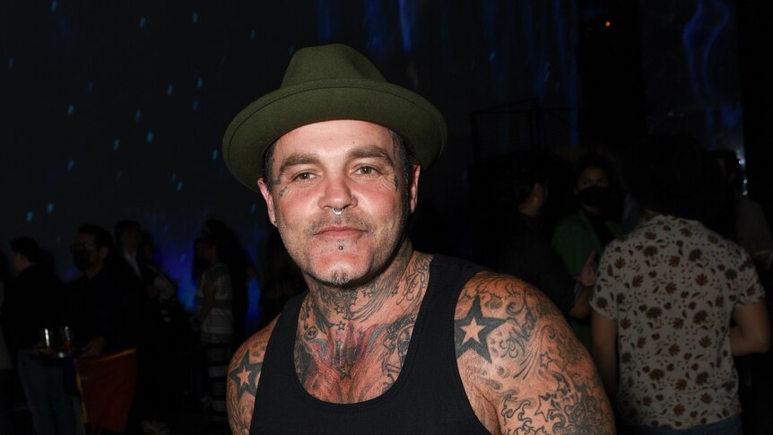 A man with tattoos on his arms, chest and neck poses for a photo, wearing a black tank top and a hat