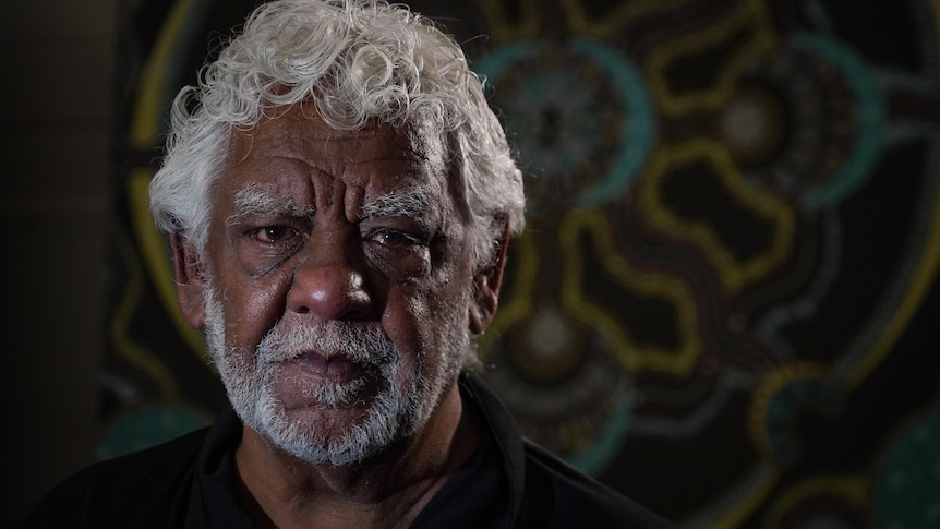 A phot of an Indigenous man looking directly at the camera with Indigenous mural in background.