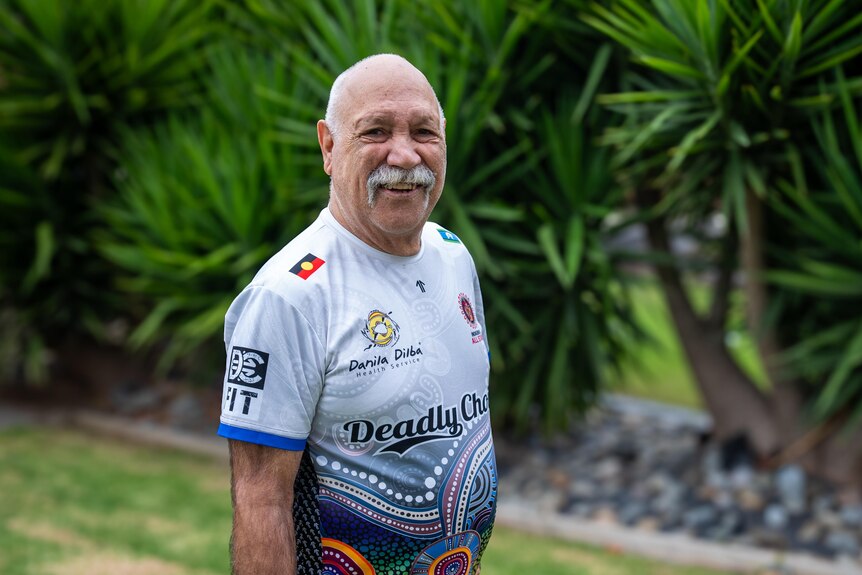 A photo showing an elderly man wearing an AFL shirt standing in front of plants. 