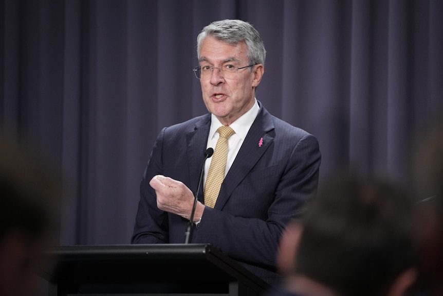 A medium shot of a man in a suit and tie standing and speaking into a microphone at a lecturn. He holds up one clenched hand