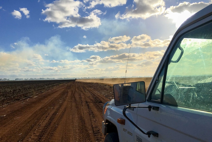 The passenger side of a ute in foreground with dust kicking up behind a vehicle on dirt road in the distance