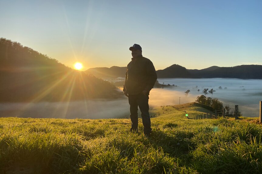 A man stand on a hill with fog in the background below and the sun creeping up behind hills in the distance.