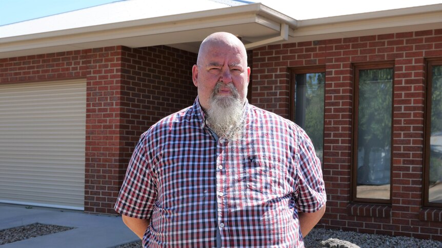 A bald man with a long grey beard, red and blue plaid shirt stands outside a red brick house with 2 vertical windows 