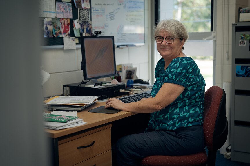 A woman with gray hair sitting at a computer in a university campus office. She's turned around in her chair and smiling.