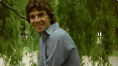 An old photograph of a young man with olive skin, floppy brown hair and a beaming smile, sitting under a willow tree.