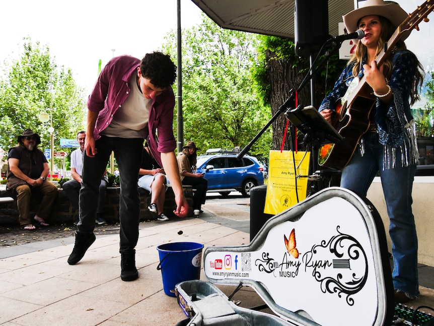 a young woman plays her guitar while someone chucked money into her guitar case