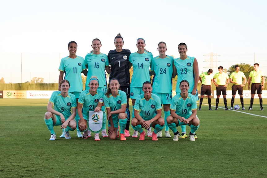 A women's soccer team wearing light blue poses for a team photo before a match with four referees in the background