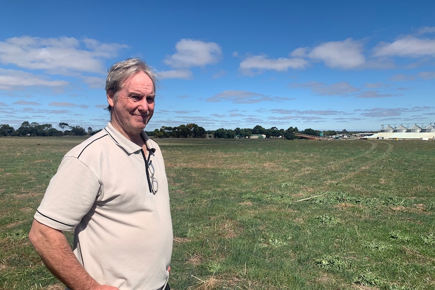 A man in a grey polo shirt stands in a green field under a blue sky.