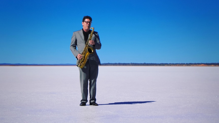 James Valentine stands on a salt flat with a blue sky in a grey suit and plays saxophone.