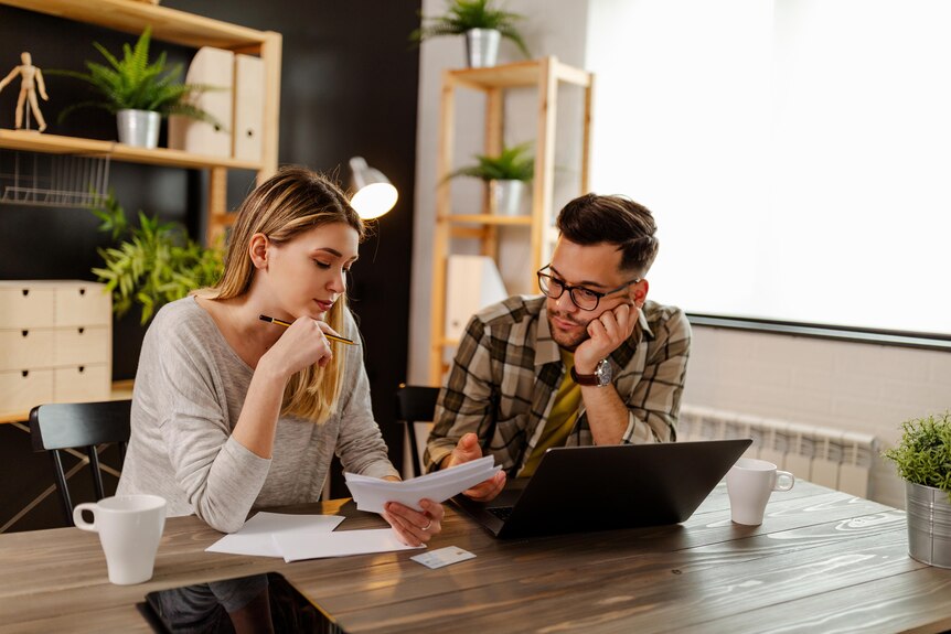 A woman and man look over finances at their laptop while sitting at a dining table.