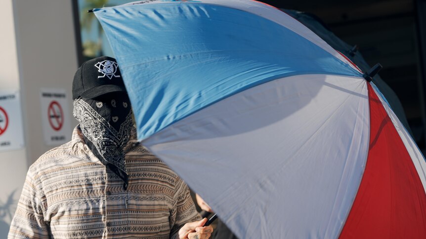 Man with bandana covering face holds up an umbrella outside court 