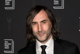 A middle-aged white man with chin-length dark hair, dark stubble and a smirk wear a suit and tie