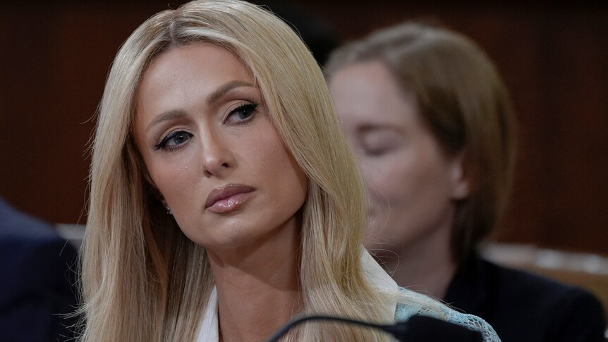 Paris Hilton with long blonde hair, serious expression on her face, tilting head to one side, wearing a pale blue suit jacket