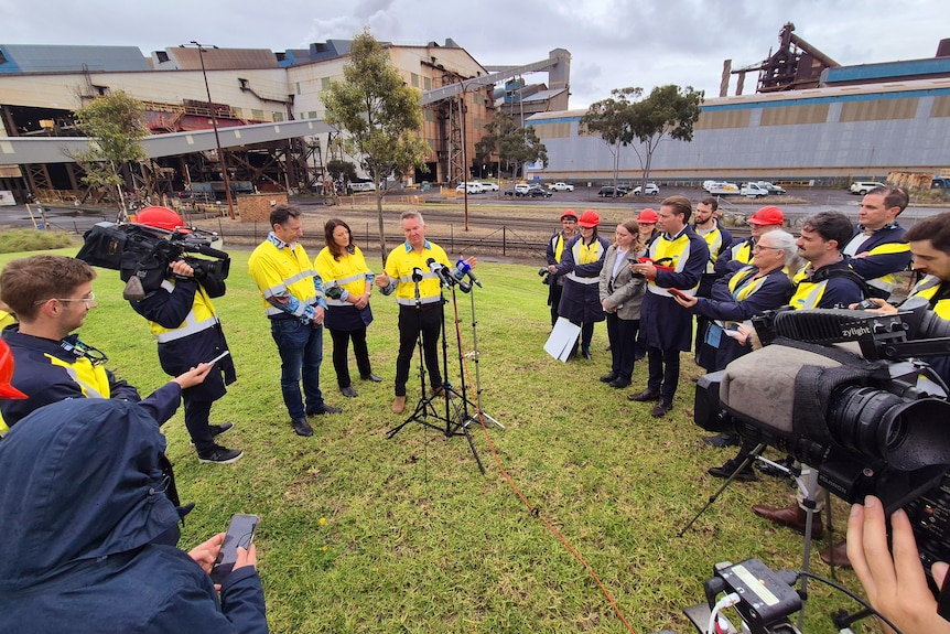 Chris Bowen addresses a large pack of media with microphones in front of him and steelworks in the background.