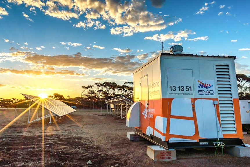 A large shed on raised tinder blocks with Western Power branding, in front of solar panels with the sun setting in background.