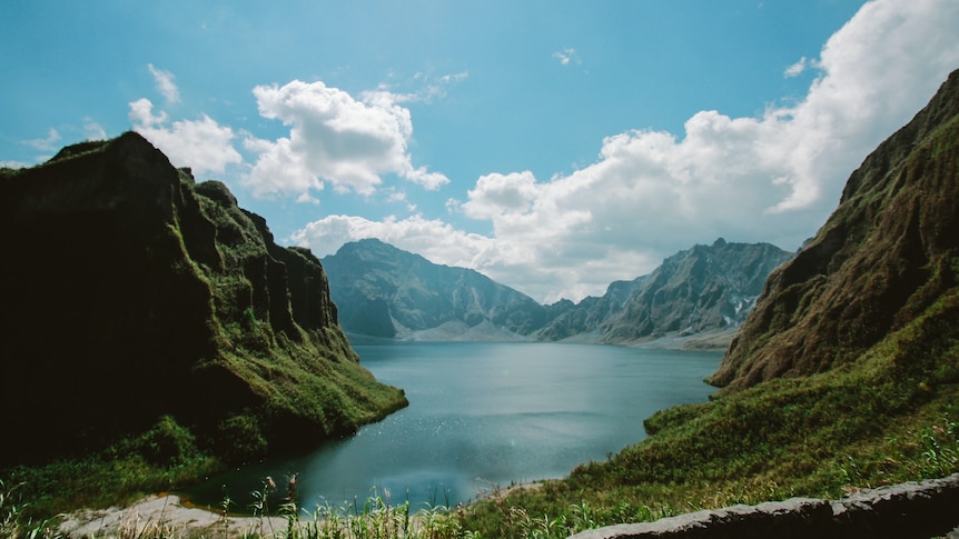 View of a large lake formed in a volcanic crater, with craggy, verdant mountains lining its shores. 