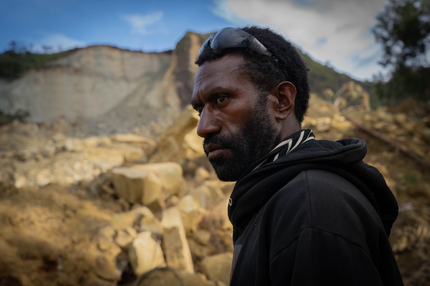 A young Papua New Guinea man wearing a black hoodie and sunglasses looks sombrely out over a large pile of rocks.