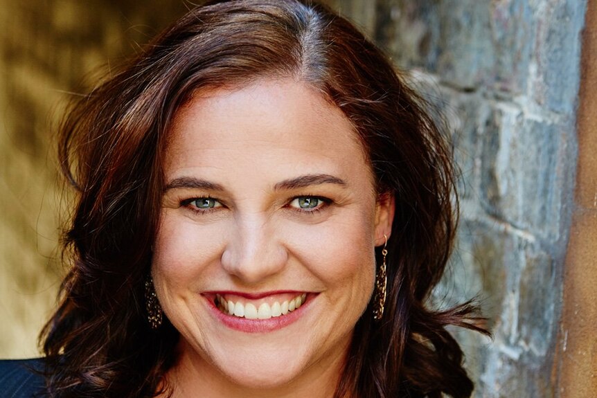 Nicola Moriarty smiling broadly wearing a low cut floral shirt and suit jacket, brown hair, blueish eyes, earrings, brick wall