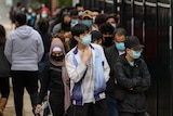 people wearing masks stand in a queue along a fenceline  